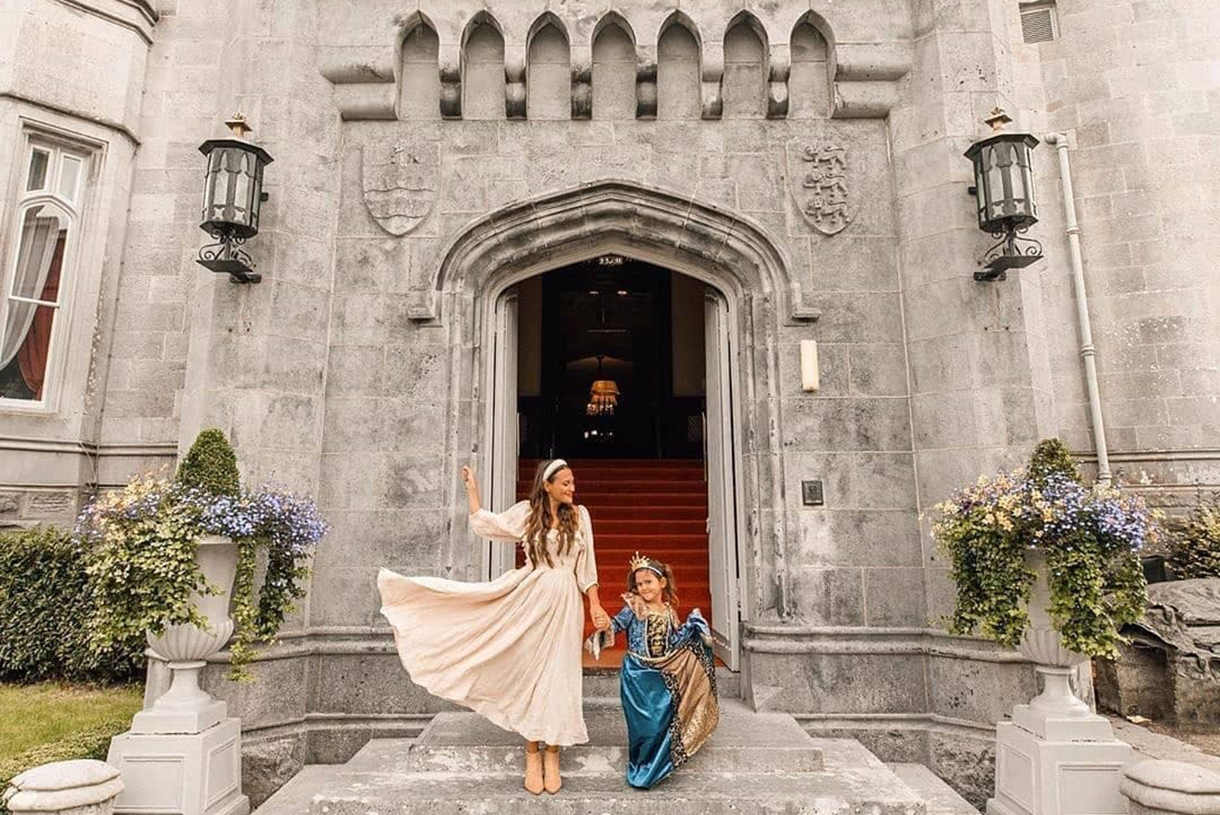 Princess and young girl dressed in royal attire standing in front of Dromoland Castle front steps. The young girl has a crown and blue gown on. 